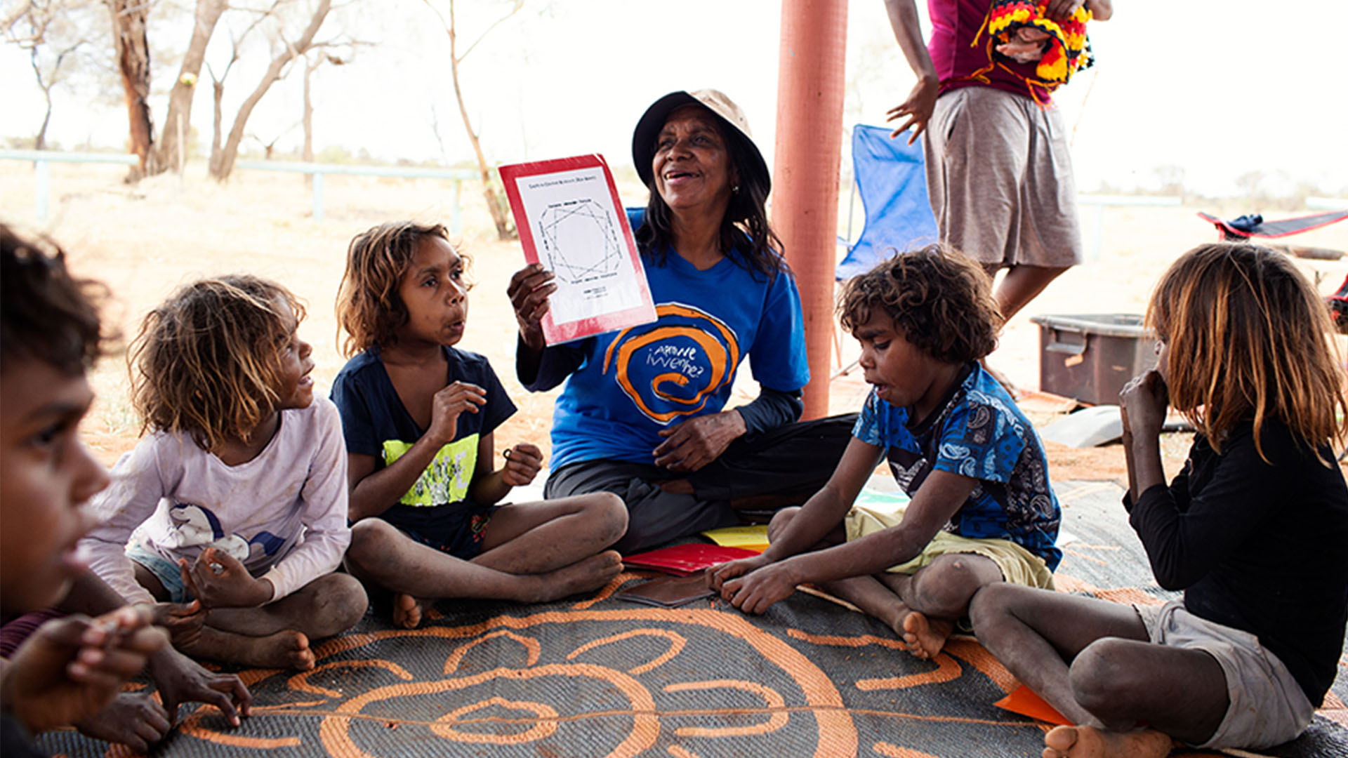 An instructor at Children's Ground shows educational materials to a group of First Nations children while all sit on the ground in a circle.