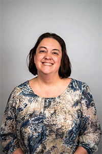 Portrait of a smiling woman with short dark hair wearing a blue and beige patterned blouse, facing the camera.