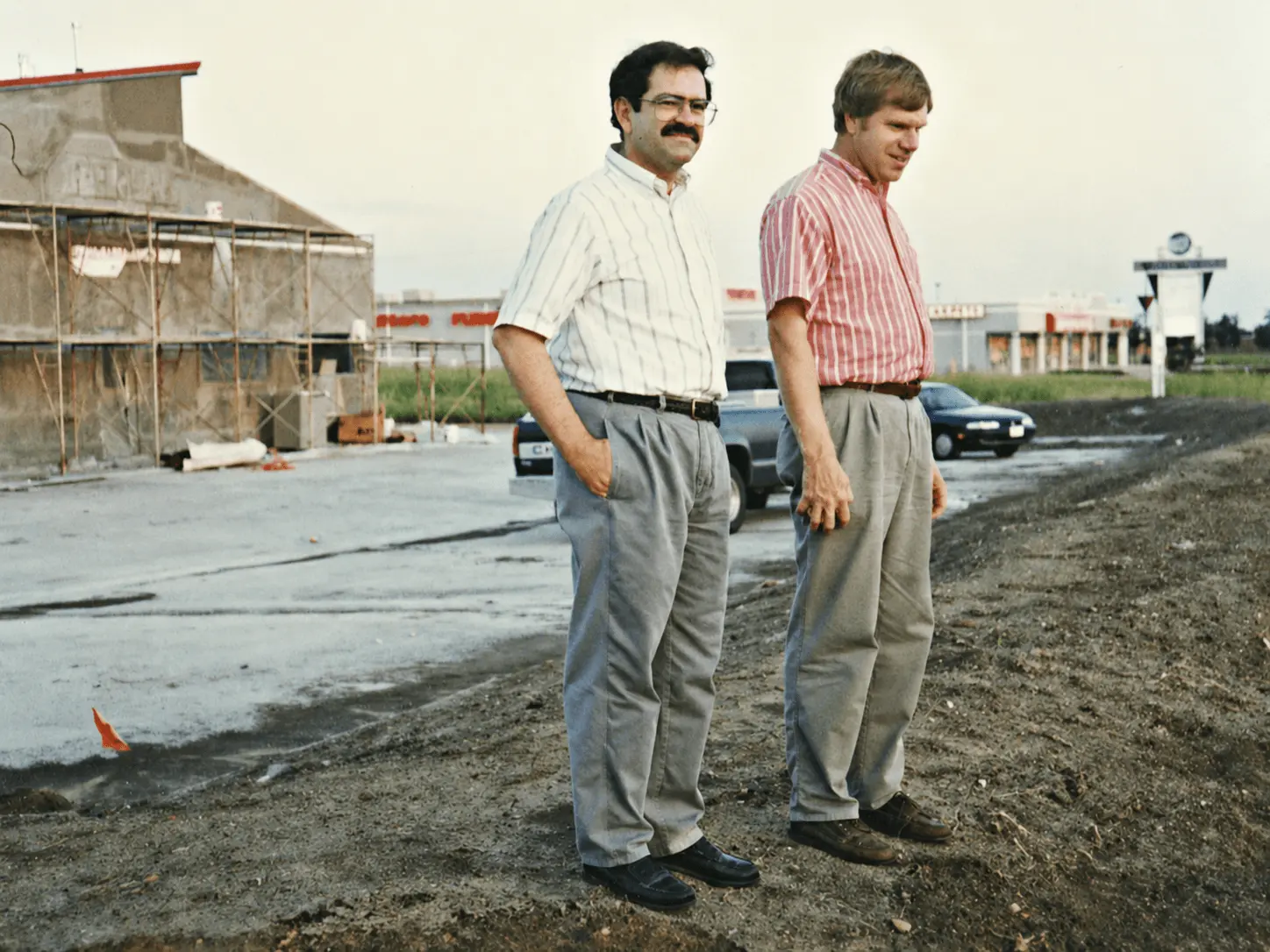 Two men in early-1990s clothing stand in a dirt lot beside the original Point of Rental store under construction in Grand Prairie, Texas, with a strip mall and parked cars in the background.